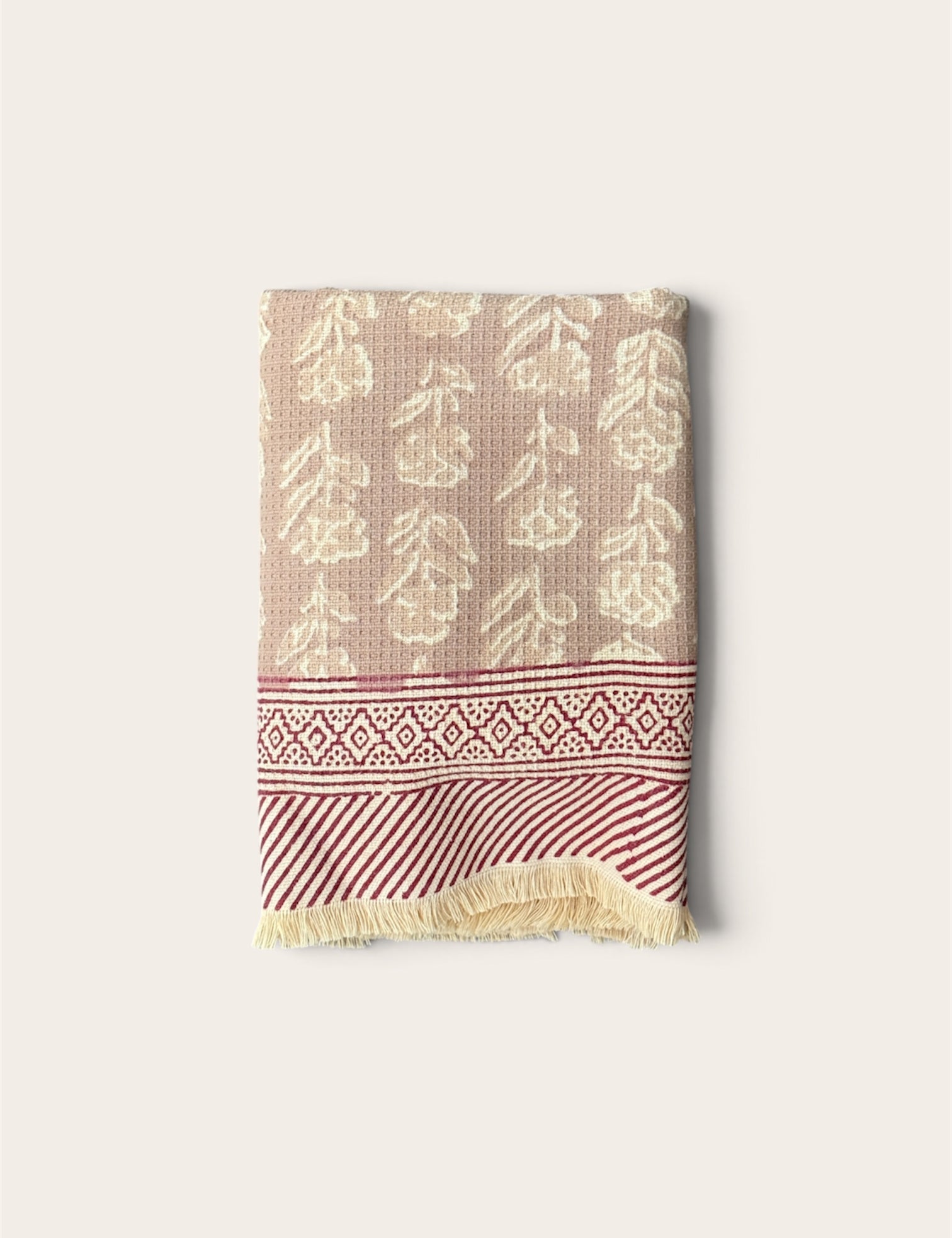 A folded woven textile, a bath towel featuring a beige and cream floral pattern on the upper section, a red and cream geometric border, and a red and cream striped section with cream fringe at the bottom, set against a solid off-white background.