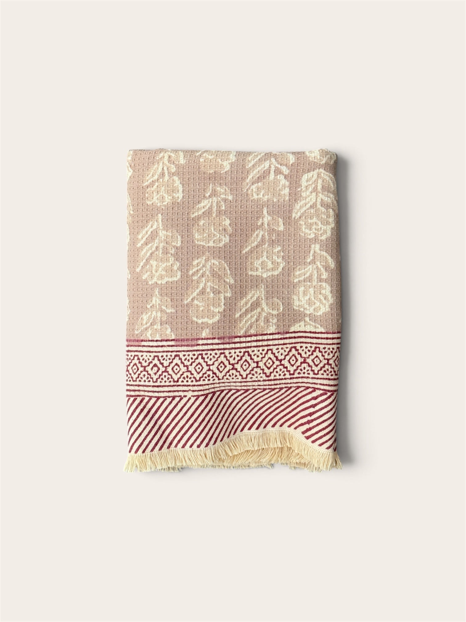 A folded woven textile, a bath towel featuring a beige and cream floral pattern on the upper section, a red and cream geometric border, and a red and cream striped section with cream fringe at the bottom, set against a solid off-white background.