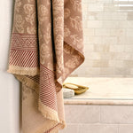 A beige and brown patterned cotton bath towel with fringed edges hangs over the side of a tiled bathroom tub, next to a gold soap dish.