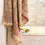A beige and brown patterned cotton bath towel with fringed edges hangs over the side of a tiled bathroom tub, next to a gold soap dish.
