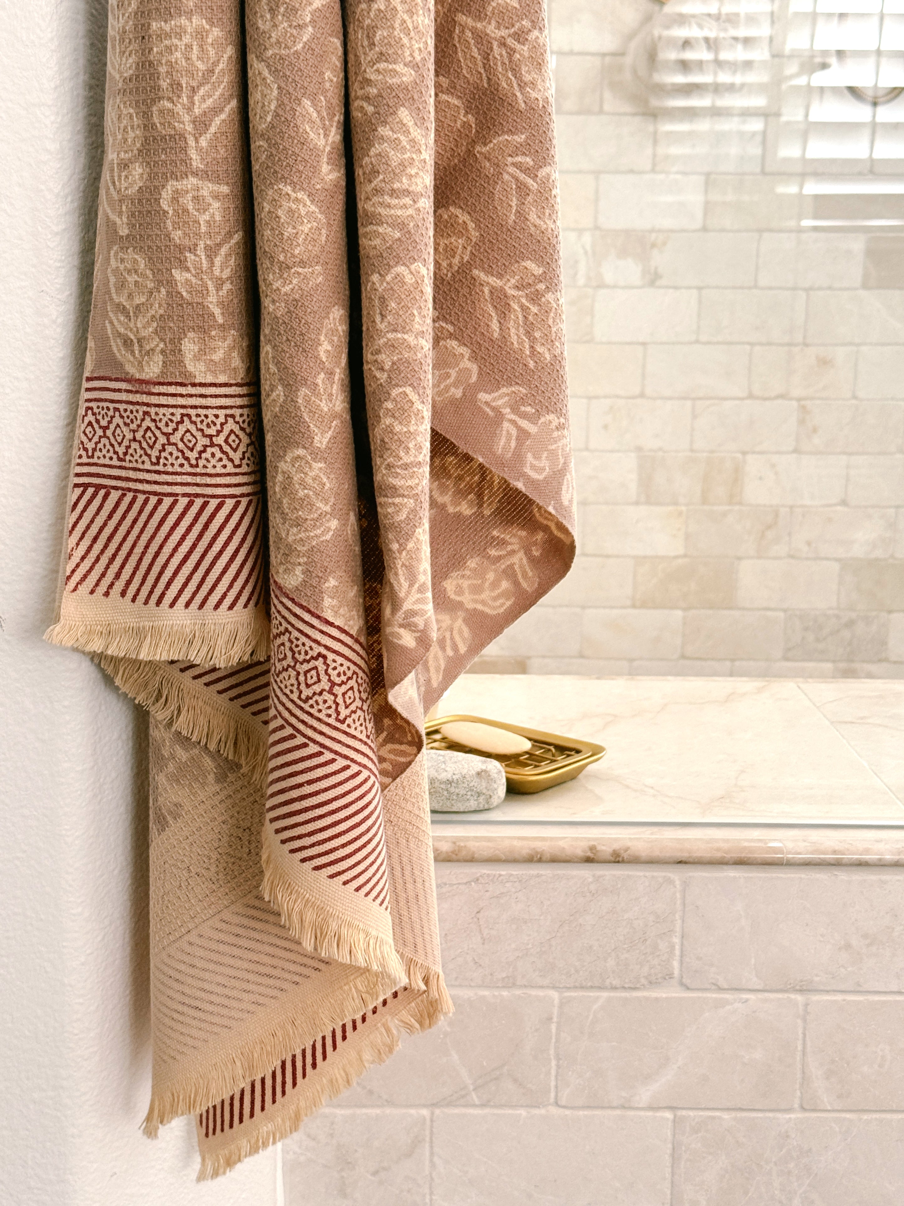 A beige and brown patterned cotton bath towel with fringed edges hangs over the side of a tiled bathroom tub, next to a gold soap dish.