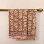 Brown woven towels with a cream leaf pattern and a decorative border, hanging from a brass towel bar against a textured off-white wall.
