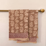 Brown woven towels with a cream leaf pattern and a decorative border, hanging from a brass towel bar against a textured off-white wall.
