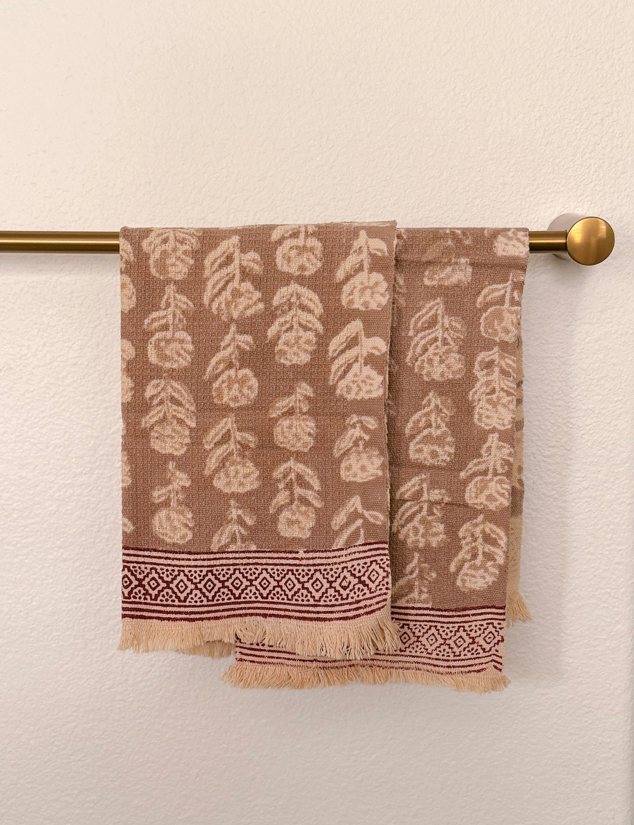 Brown woven towels with a cream leaf pattern and a decorative border, hanging from a brass towel bar against a textured off-white wall.