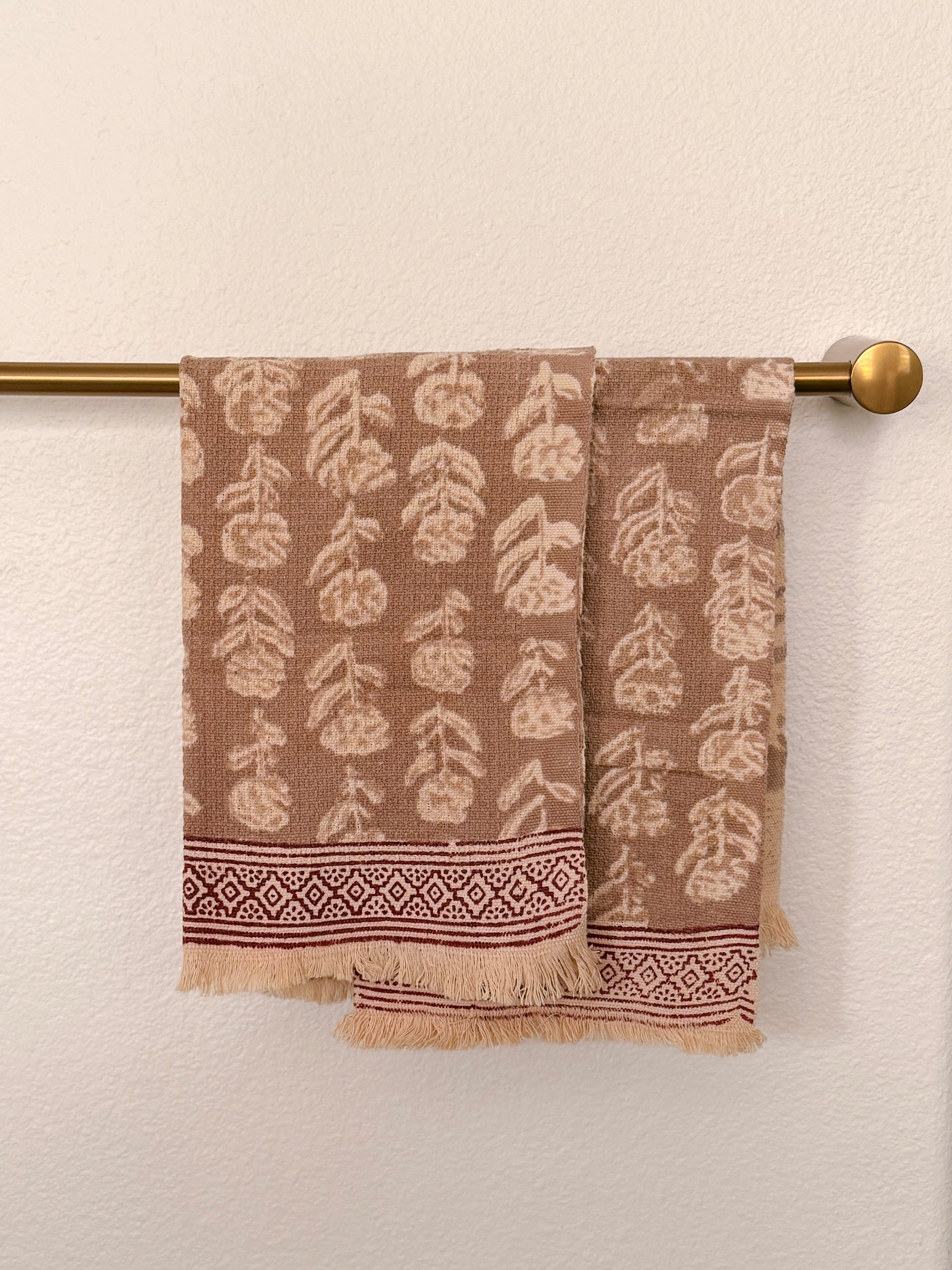 Brown woven towels with a cream leaf pattern and a decorative border, hanging from a brass towel bar against a textured off-white wall.