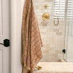 A beige and rose pink floral patterned cotton bath towel hangs from a brass hook on a white door frame in a modern bathroom with beige tile walls and gold fixtures.