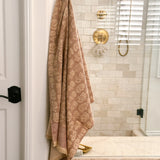 A beige and rose pink floral patterned cotton bath towel hangs from a brass hook on a white door frame in a modern bathroom with beige tile walls and gold fixtures.