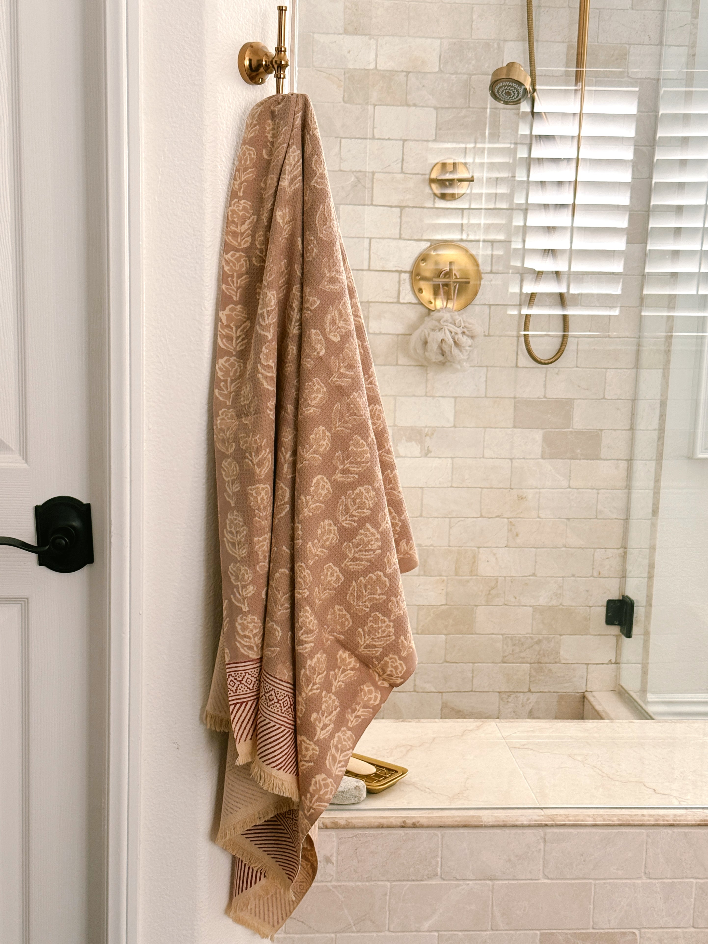 A beige and rose pink floral patterned cotton bath towel hangs from a brass hook on a white door frame in a modern bathroom with beige tile walls and gold fixtures.