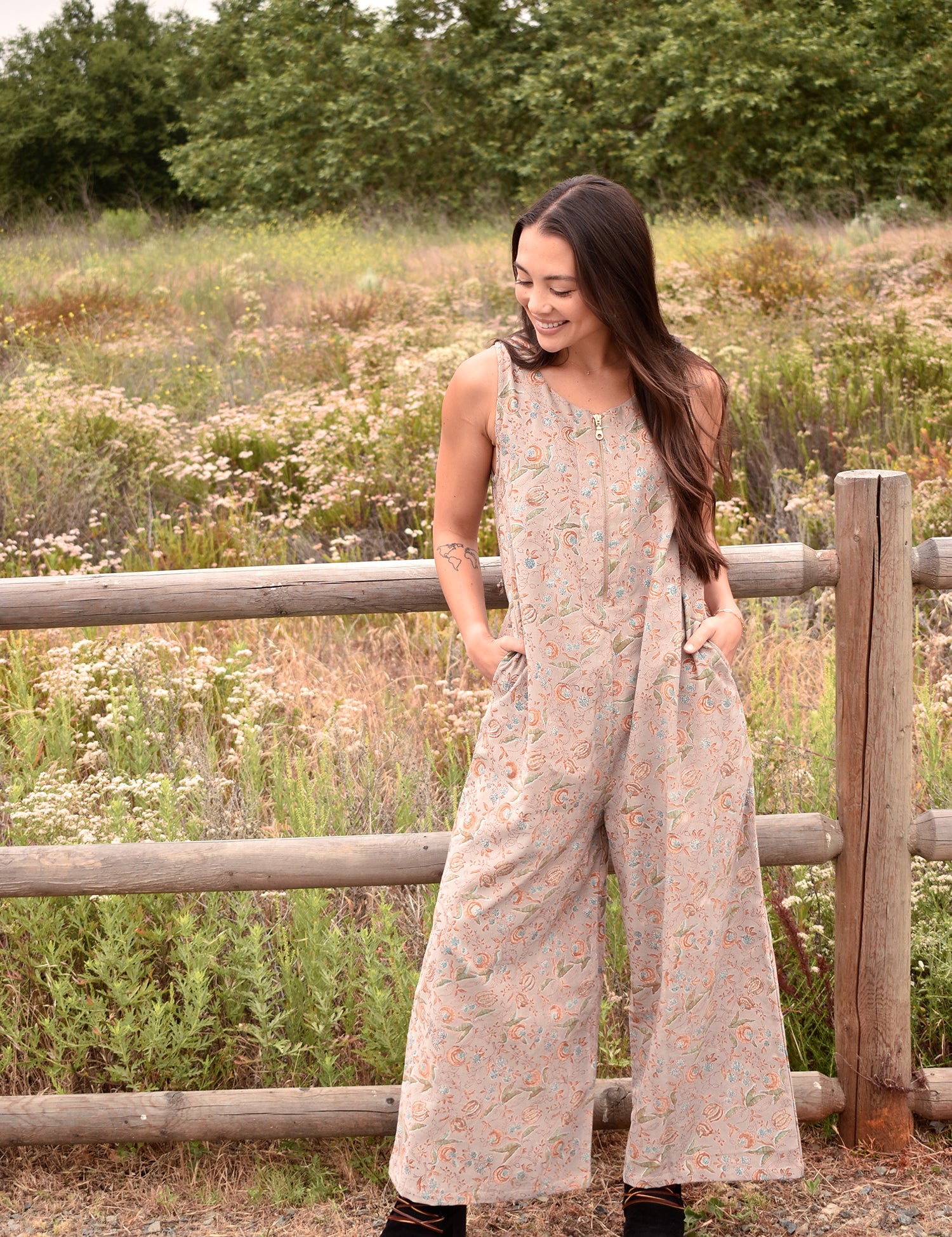 Woman wearing a floral printed gray jumpsuit with boots, standing in front of a fence and trees and bushes