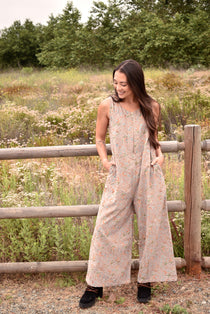 Woman wearing a floral printed gray jumpsuit with boots, standing in front of a fence and trees and bushes