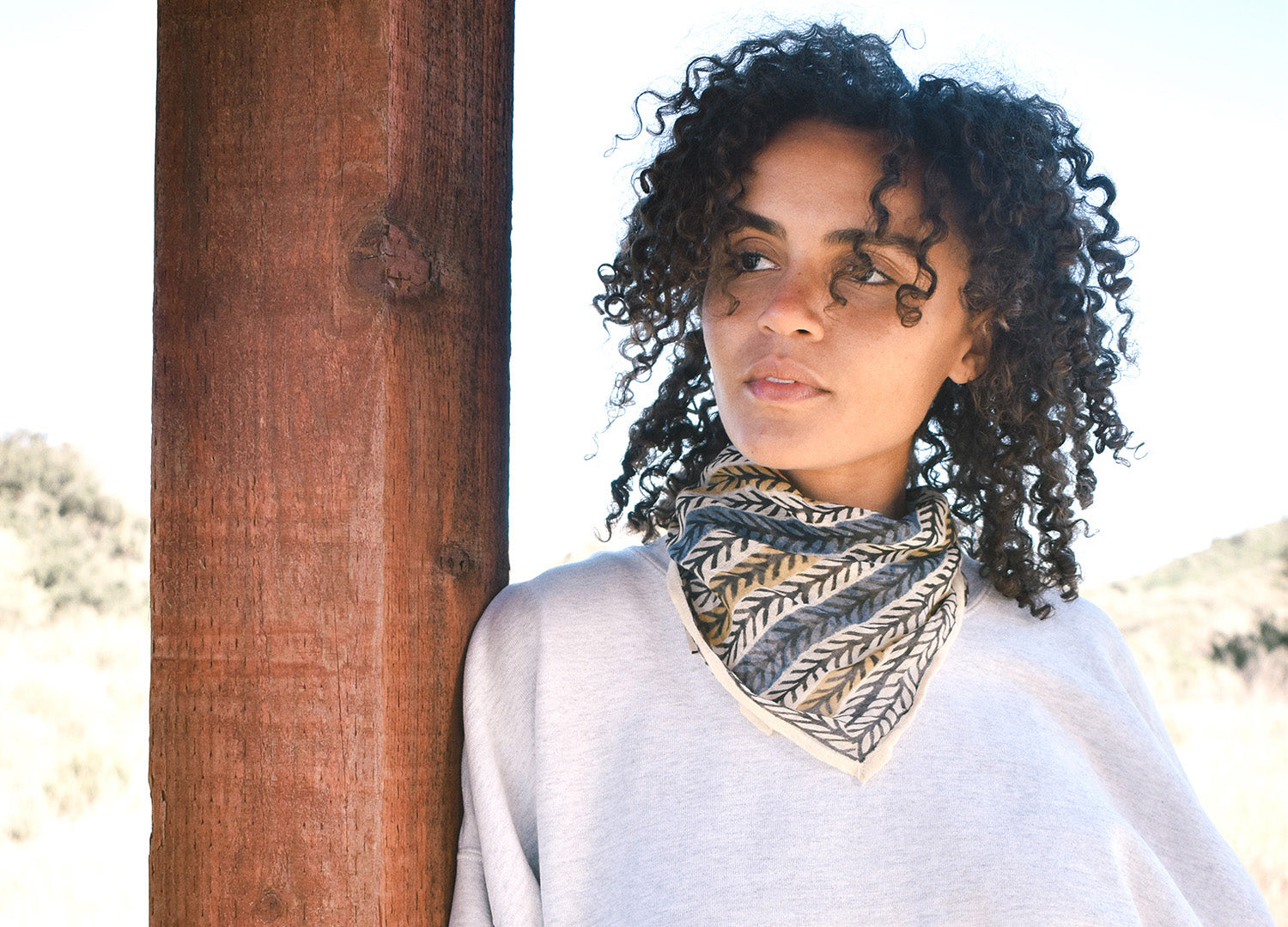 Woman wearing block print Frontier bandana tied around neck against a grey sweatshirt. Woman leaning against wooden pillar.