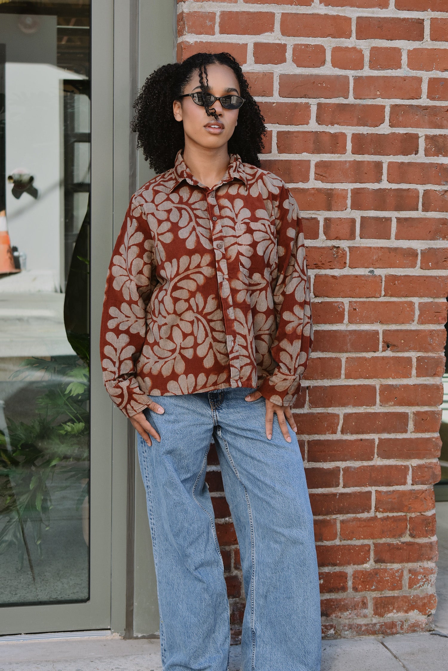 Person wearing a floral patterned corduroy shirt and jeans standing against a brick wall.