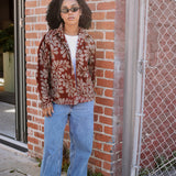 Person wearing a floral patterned shirt open as a jacket and blue jeans standing against a brick wall.