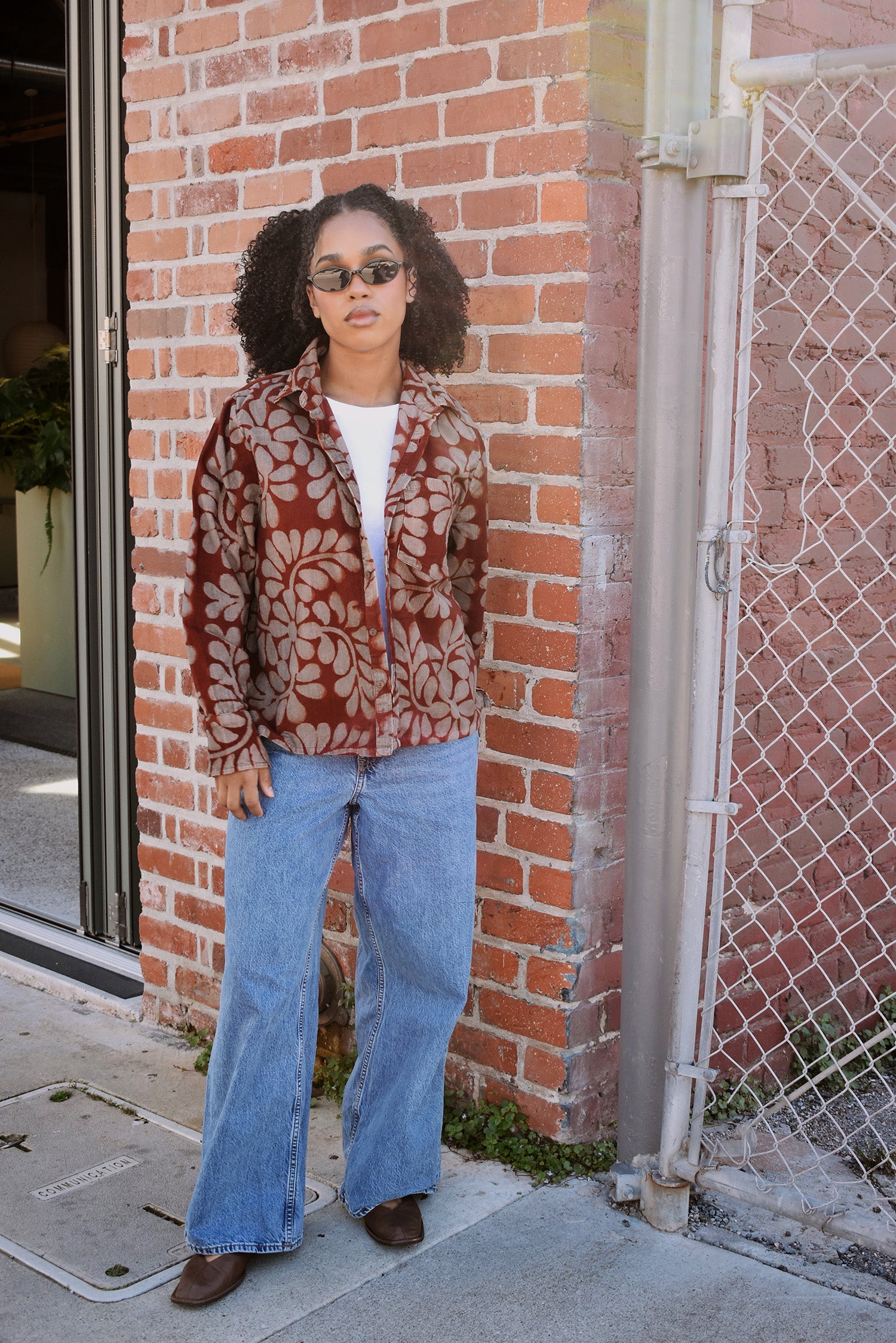 Person wearing a floral patterned shirt open as a jacket and blue jeans standing against a brick wall.