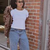 Woman wearing a white t-shirt and blue jeans leaning against a brick wall, putting on a floral printed shirt