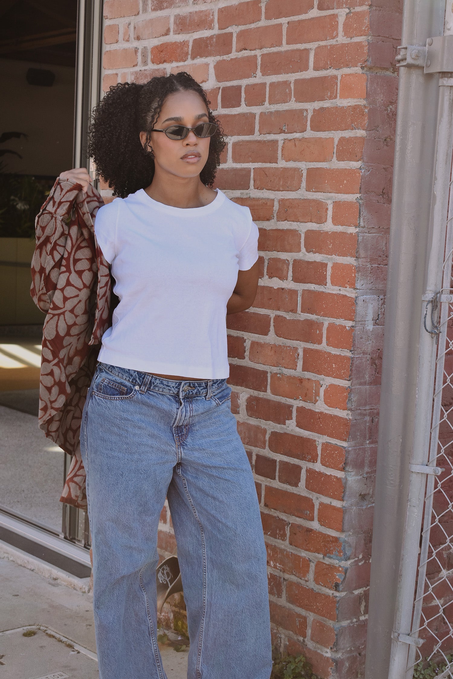 Woman wearing a white t-shirt and blue jeans leaning against a brick wall, putting on a floral printed shirt