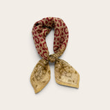 A floral and heart patterned cotton scarf tied in a knot, featuring a red floral motif and a sage floral border print, set against a plain light beige background.