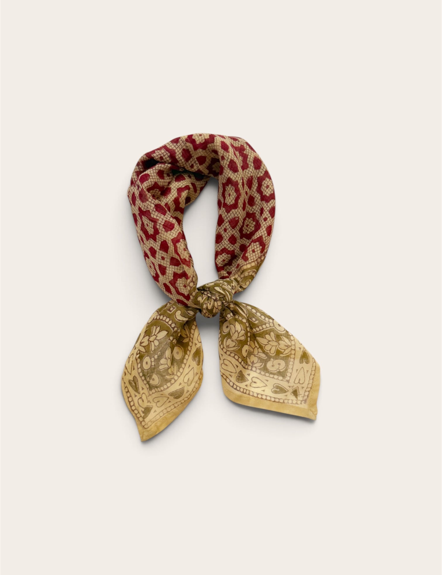 A floral and heart patterned cotton scarf tied in a knot, featuring a red floral motif and a sage floral border print, set against a plain light beige background.