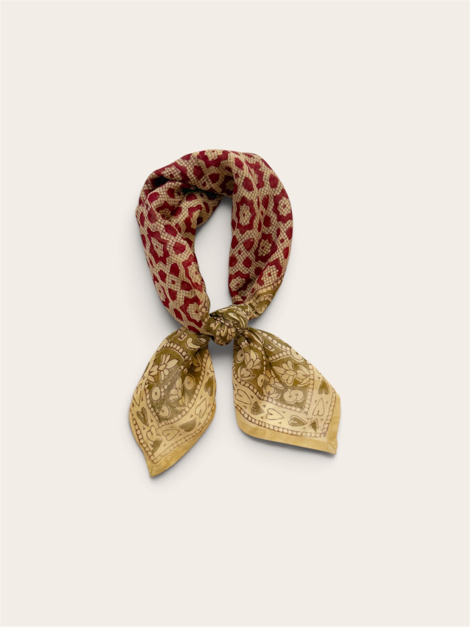 A floral and heart patterned cotton scarf tied in a knot, featuring a red floral motif and a sage floral border print, set against a plain light beige background.