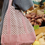Woman wearing red checkered and striped tote bag in front of potato section at grocery store
