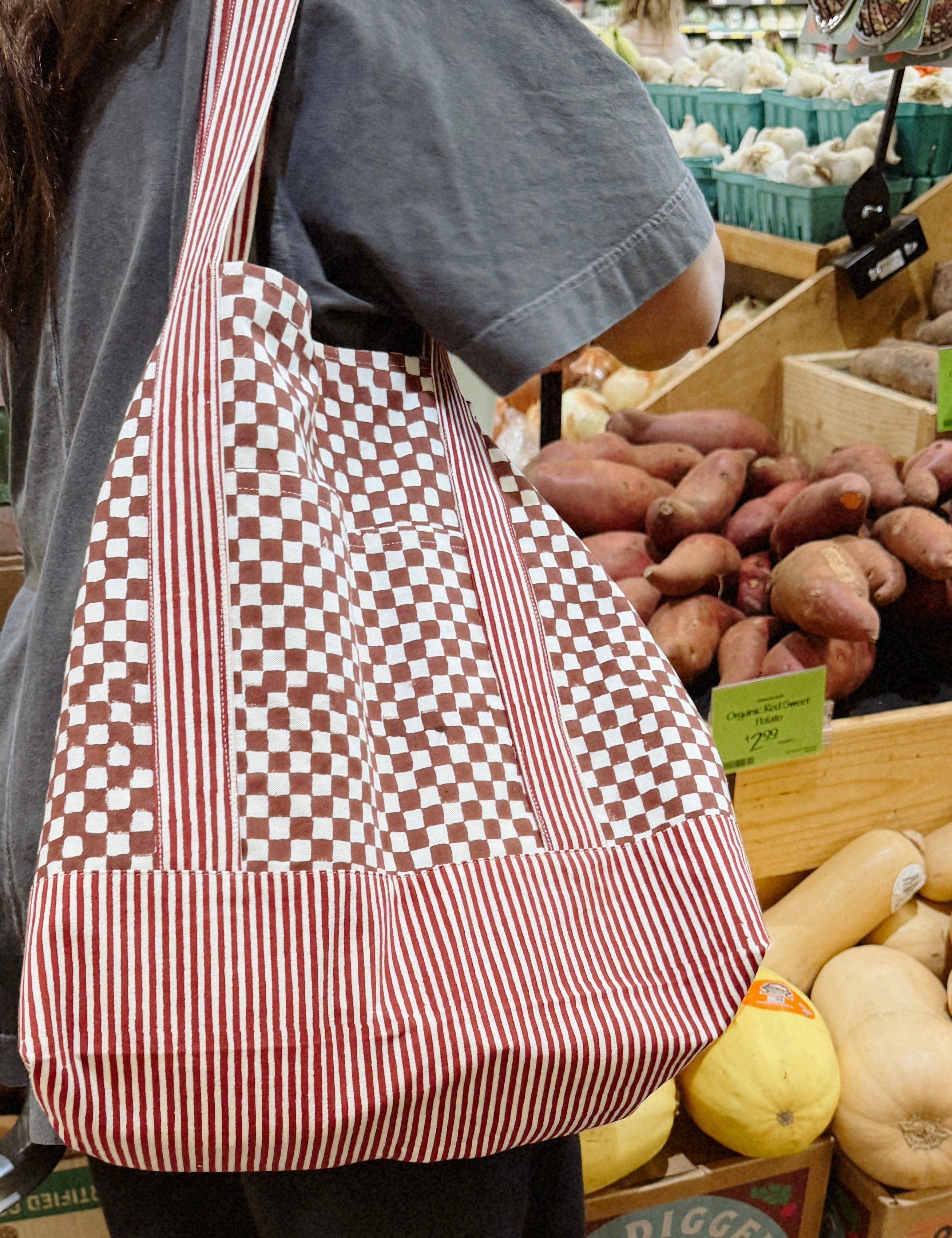 Woman wearing red checkered and striped tote bag in front of potato section at grocery store