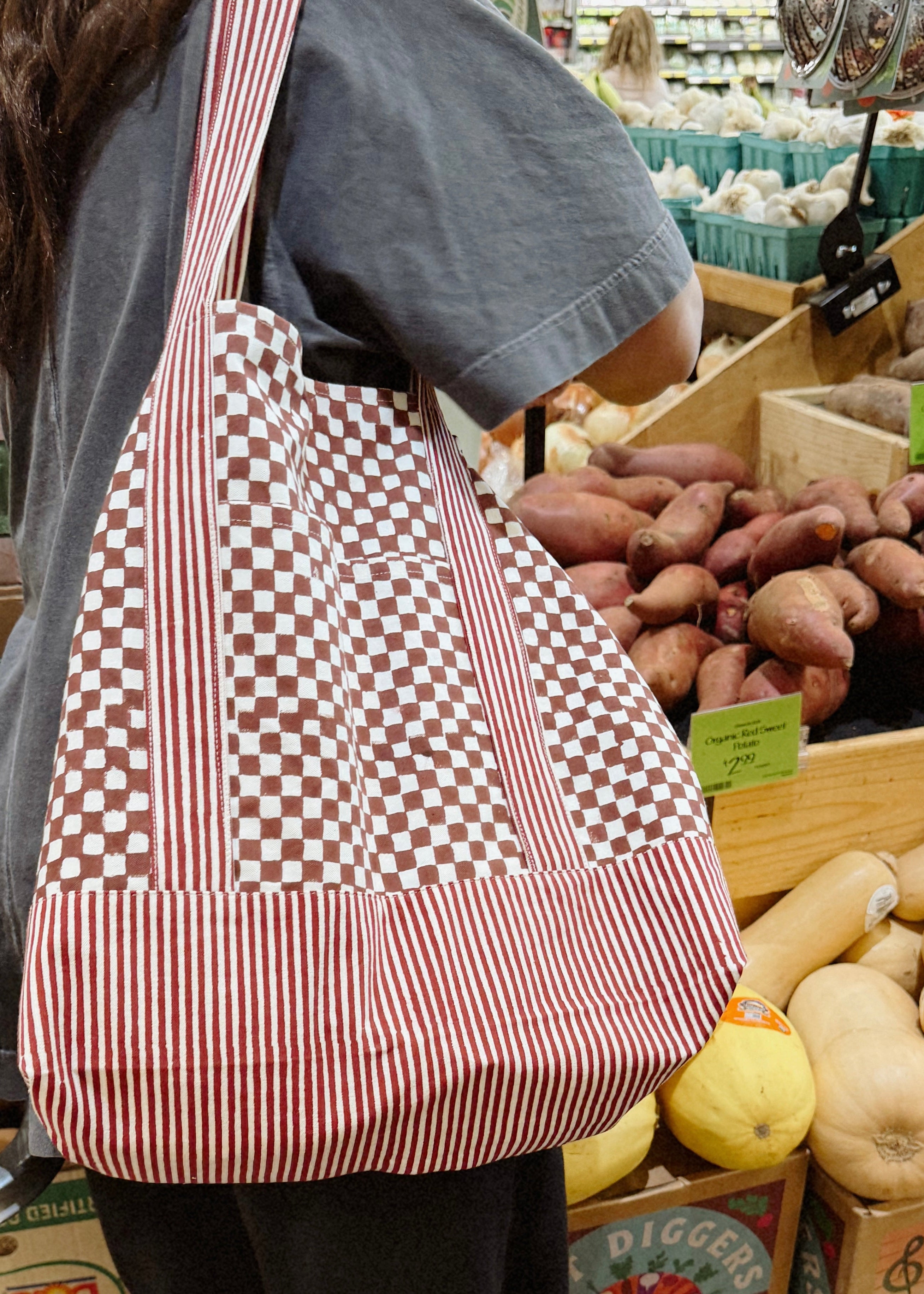 Woman wearing red checkered and striped tote bag in front of potato section at grocery store