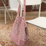 Red checkered and striped tote bag hanging on a white chair outside, rocks and leave on the ground. 