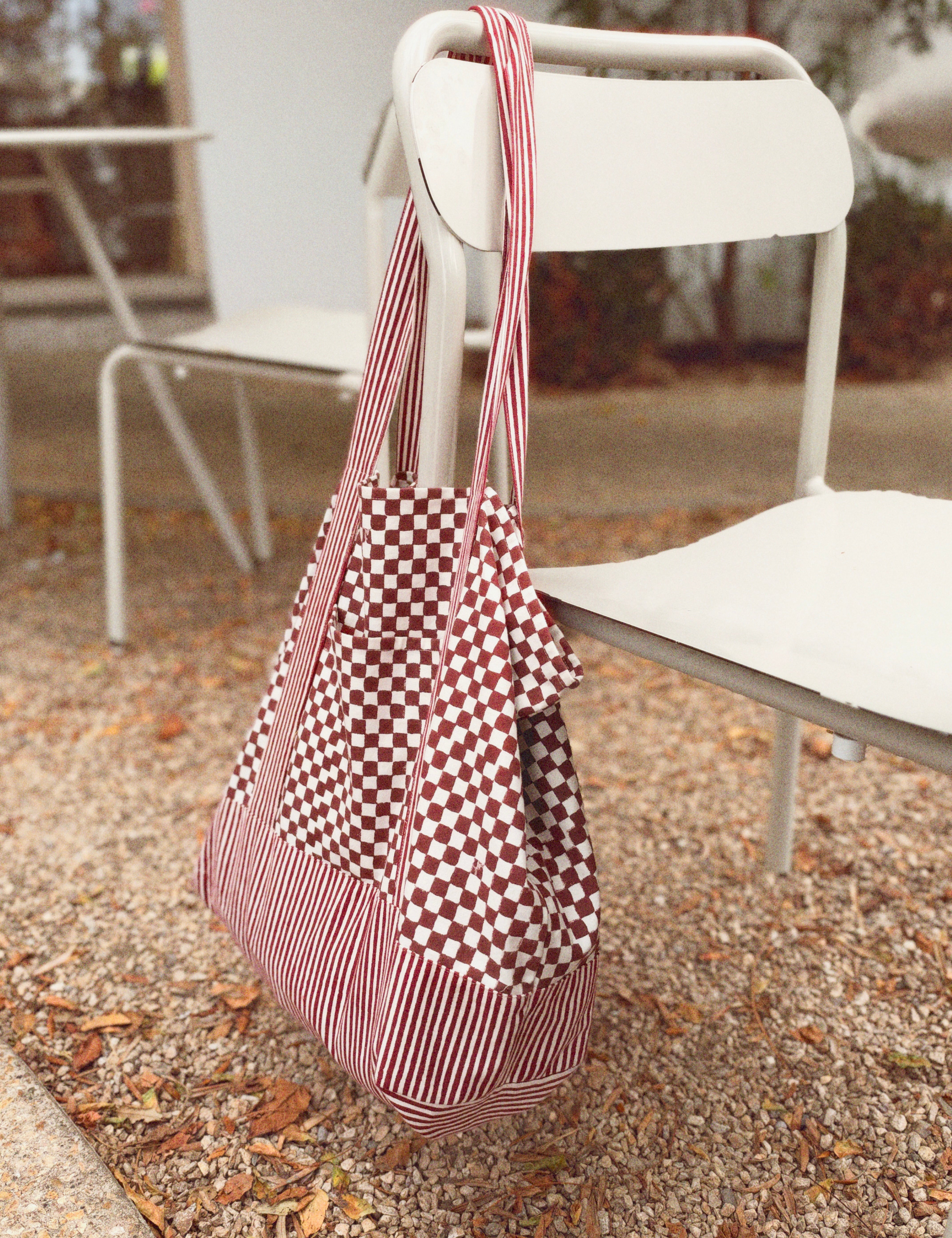 Red checkered and striped tote bag hanging on a white chair outside, rocks and leave on the ground. 