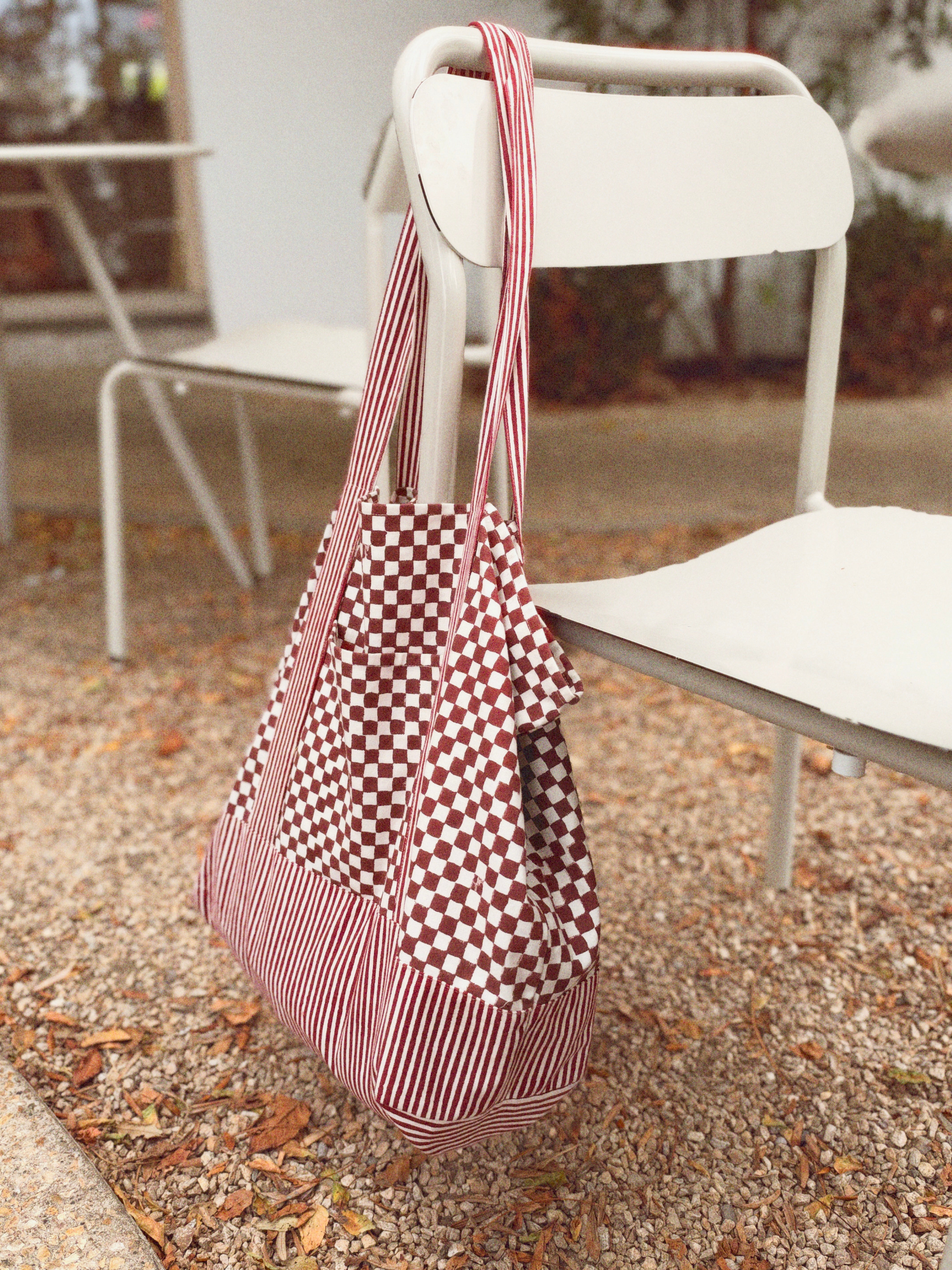 Red checkered and striped tote bag hanging on a white chair outside, rocks and leave on the ground. 