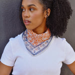 Floral printed orange and blue bandana scarf tied around a woman's neck over a white tee. Woman is standing in front of a black background. 