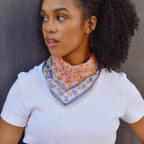 Floral printed orange and blue bandana scarf tied around a woman's neck over a white tee. Woman is standing in front of a black background. 
