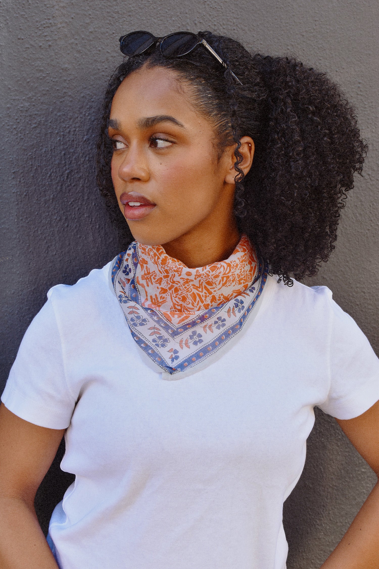 Floral printed orange and blue bandana scarf tied around a woman's neck over a white tee. Woman is standing in front of a black background. 