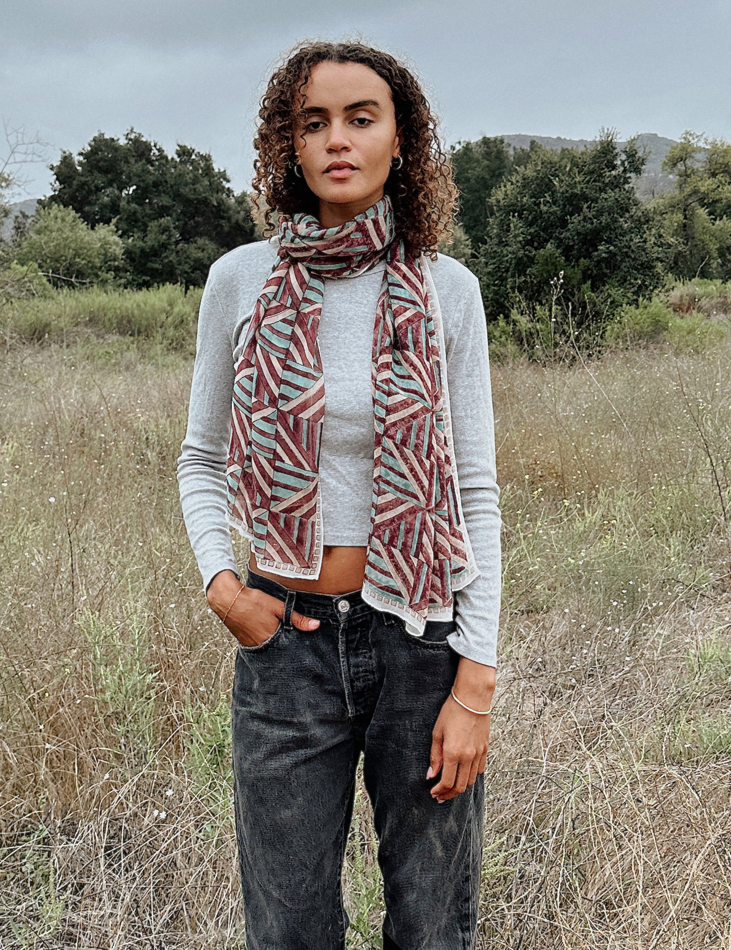 Woman wearing block printed Layla georgette scarf around the neck, paired with grey long sleeve and black jeans. Woman is standing outside.