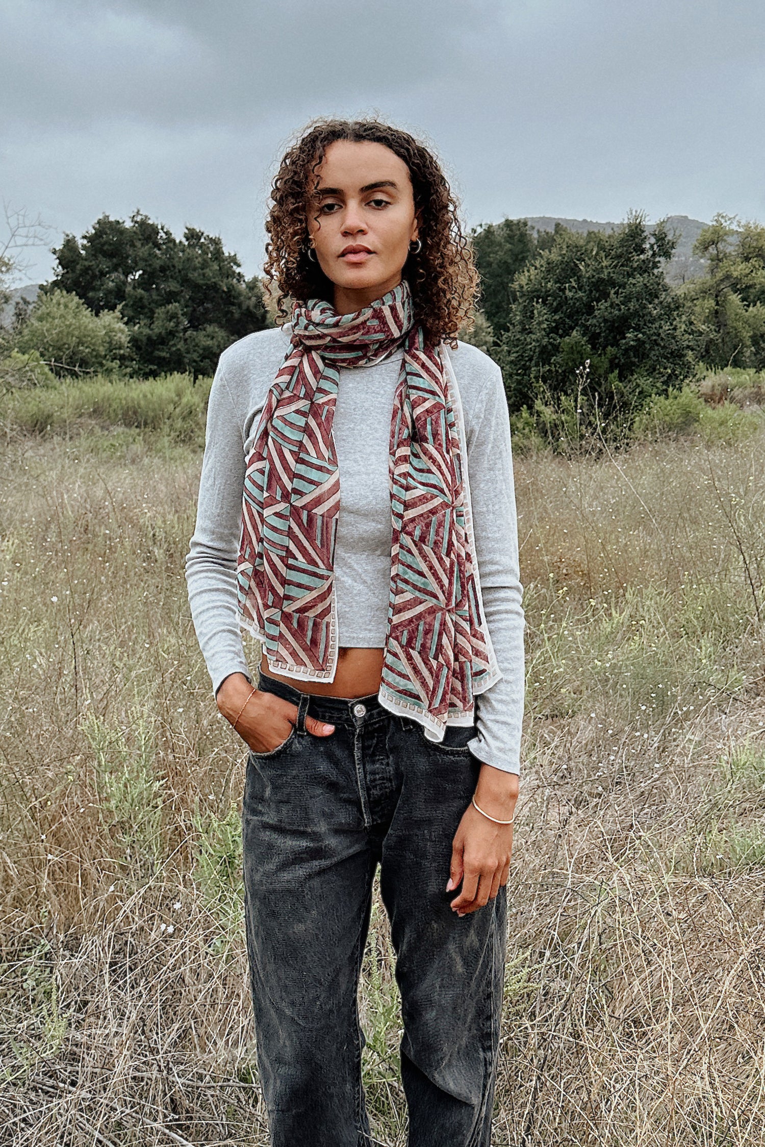 Woman wearing block printed Layla georgette scarf around the neck, paired with grey long sleeve and black jeans. Woman is standing outside.