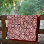 Red and pink velvet blanket with geometric floral print allover, draped over a wooden fence in a rustic setting with trees in the background. 