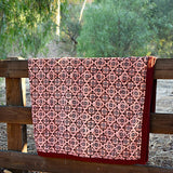 Red and pink velvet blanket with geometric floral print allover, draped over a wooden fence in a rustic setting with trees in the background. 