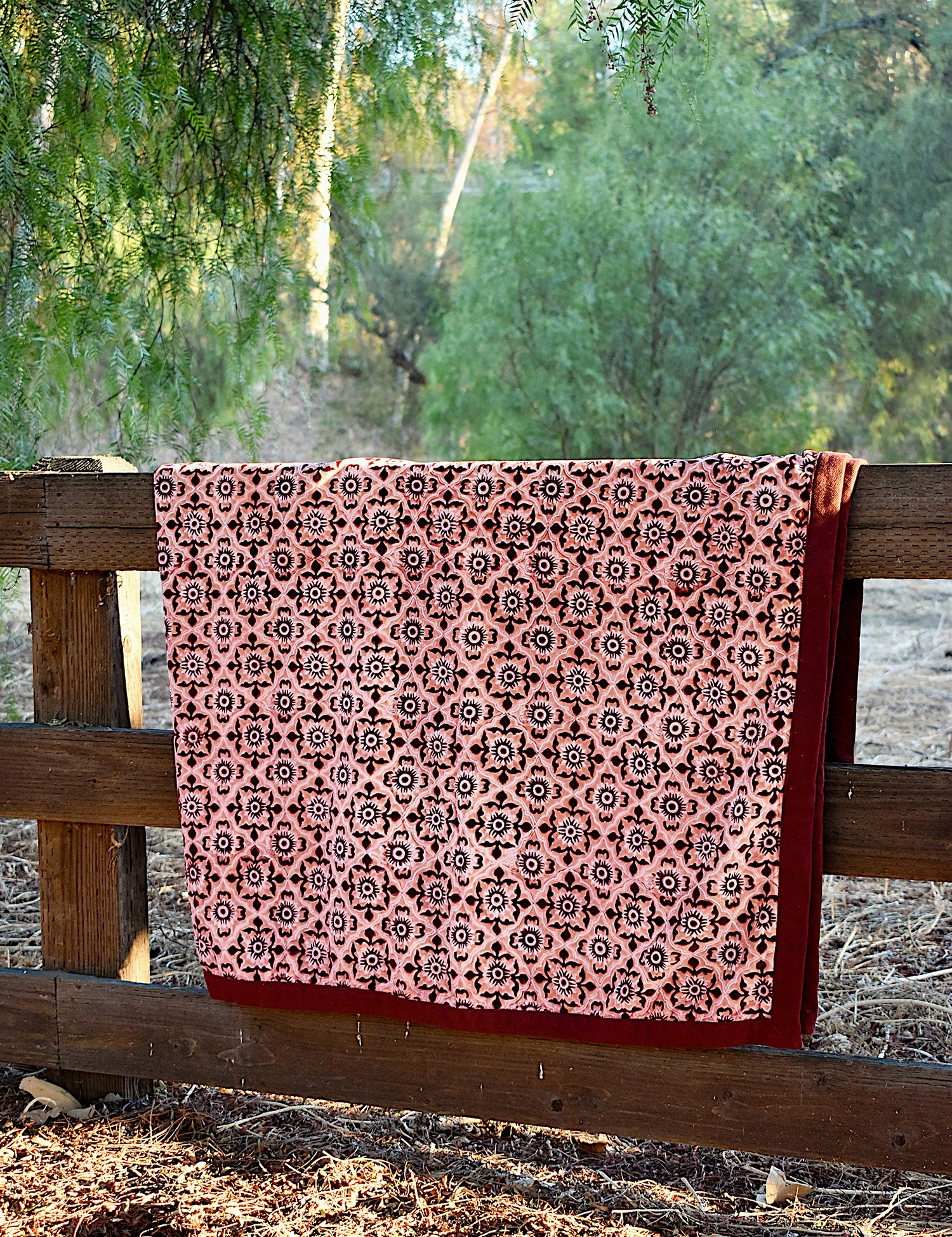 Red and pink velvet blanket with geometric floral print allover, draped over a wooden fence in a rustic setting with trees in the background. 