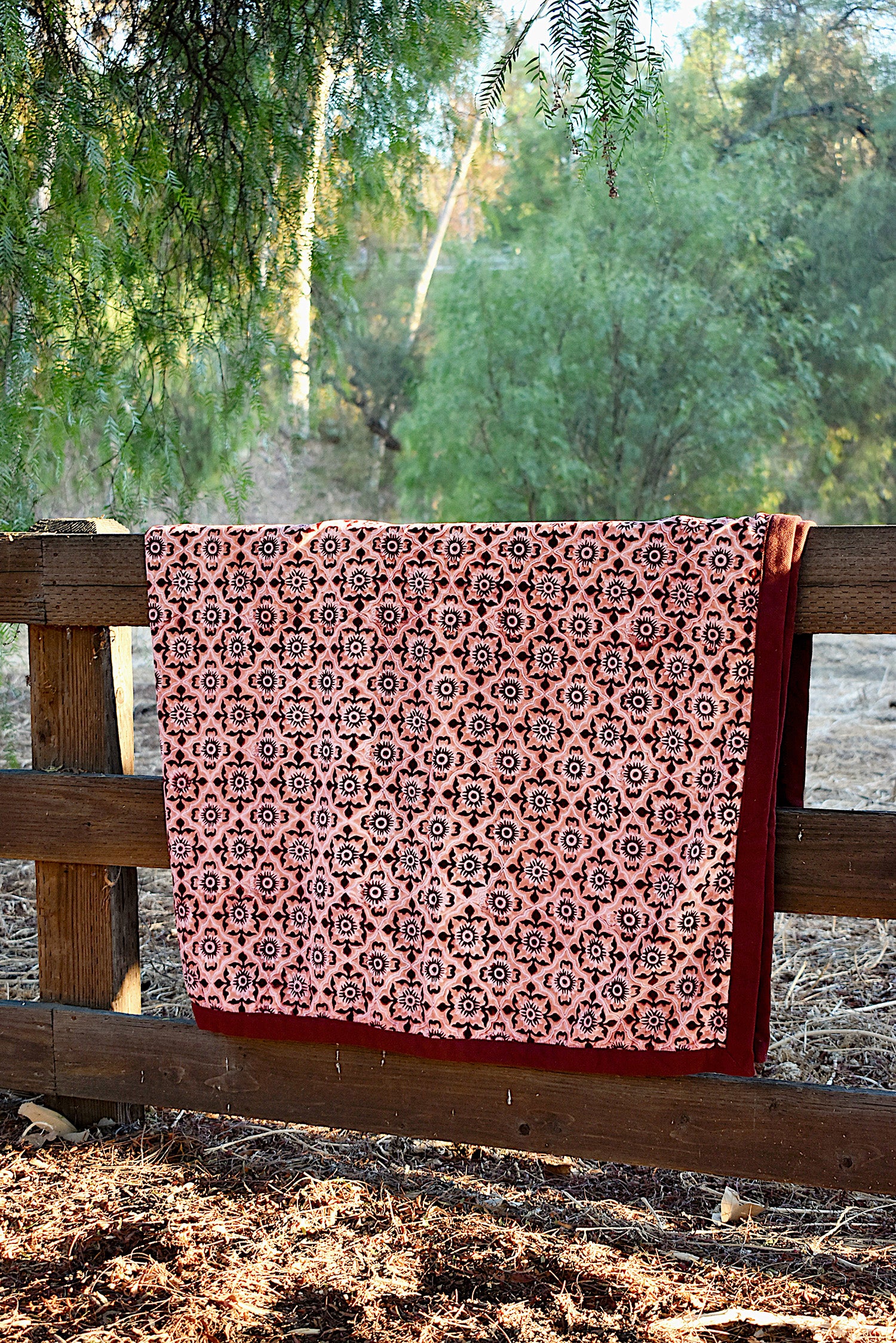 Red and pink velvet blanket with geometric floral print allover, draped over a wooden fence in a rustic setting with trees in the background. 