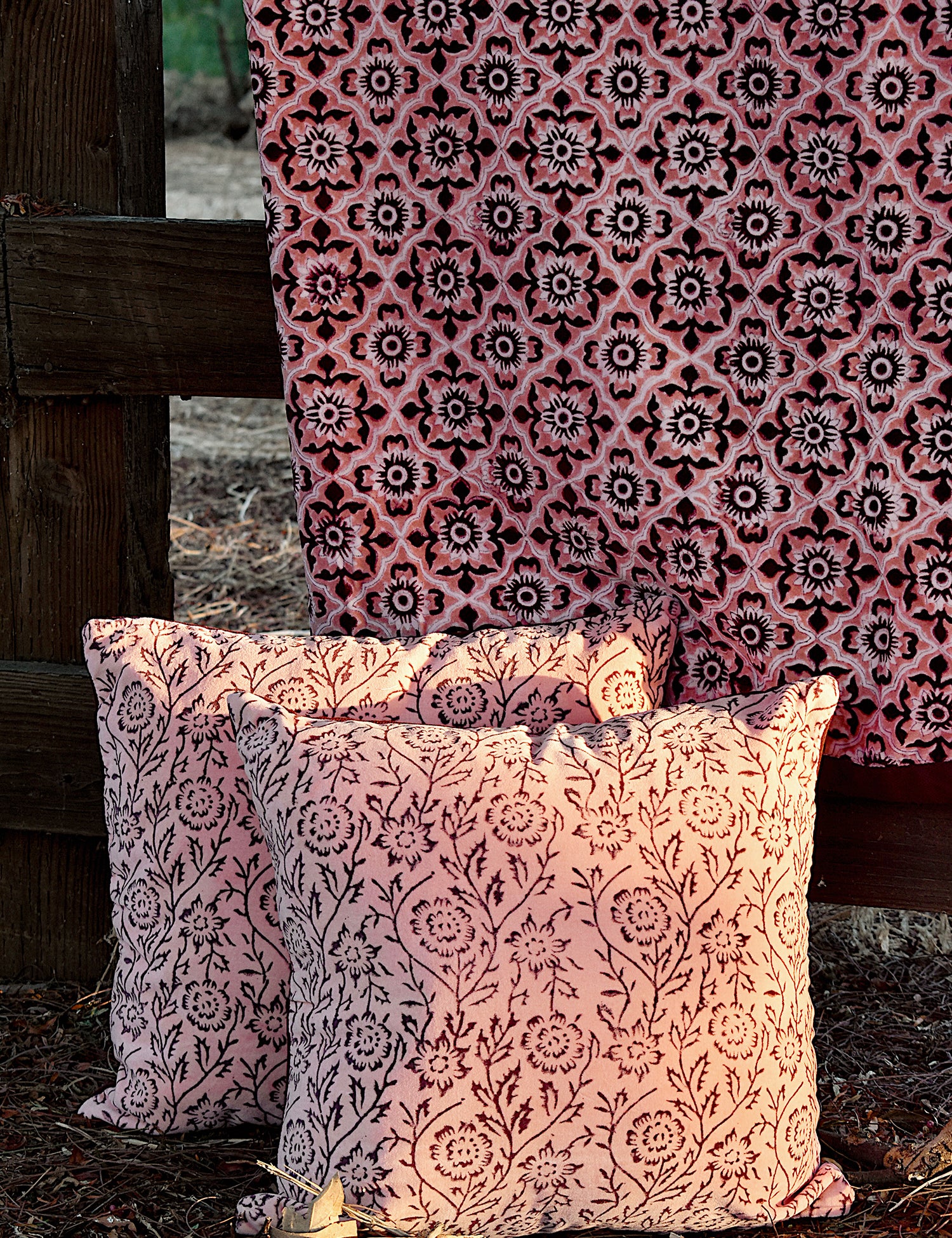 Floral printed blanket draped over a wooden fence in a rustic setting. Two pillows with different floral print sitting on ground in front of the blanket.