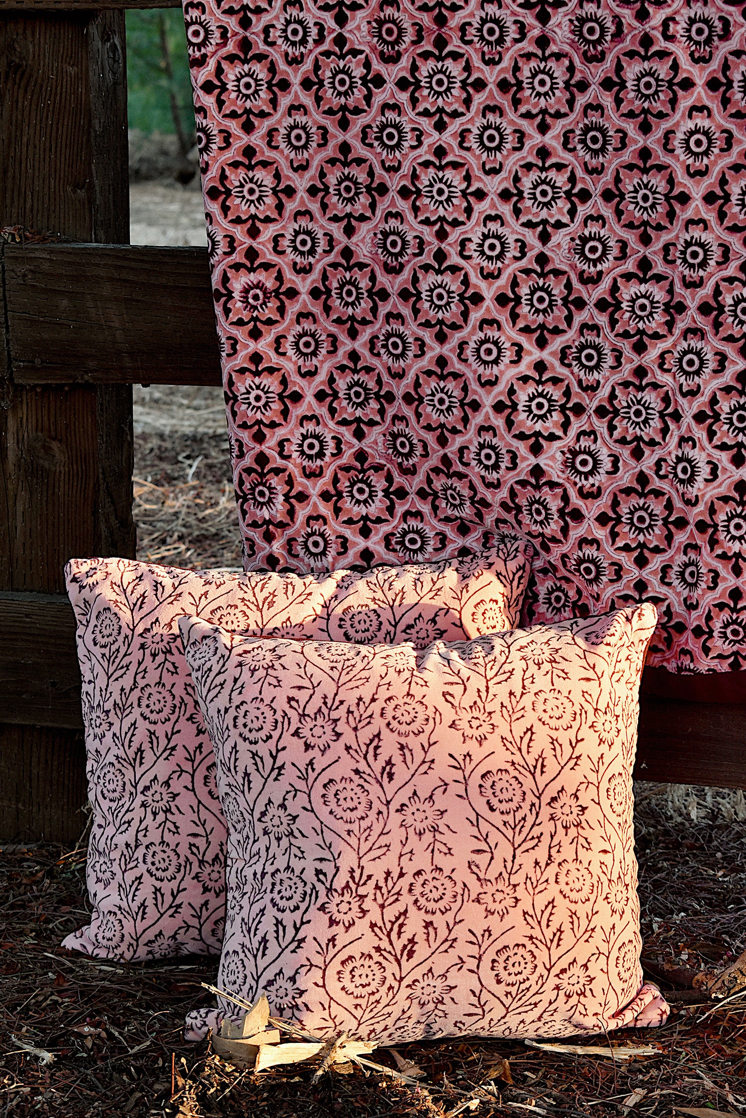 Floral printed blanket draped over a wooden fence in a rustic setting. Two pillows with different floral print sitting on ground in front of the blanket.