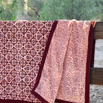 A pink and burgundy floral patterned blanket throw draped over a rustic wooden fence outdoors, with trees in the background. Blanket is open to show reversible print. 