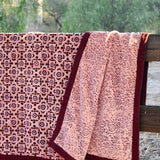 A pink and burgundy floral patterned blanket throw draped over a rustic wooden fence outdoors, with trees in the background. Blanket is open to show reversible print. 