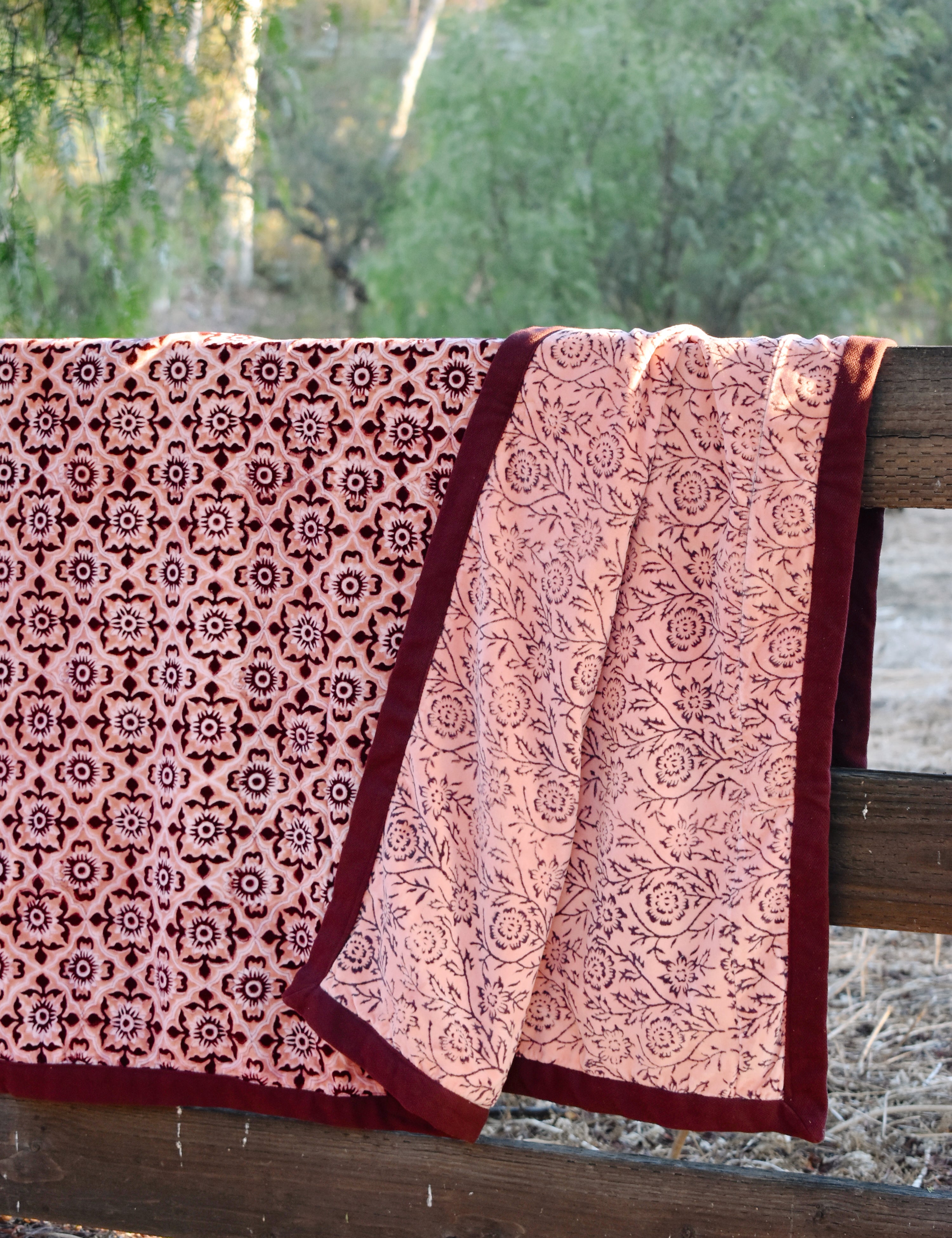A pink and burgundy floral patterned blanket throw draped over a rustic wooden fence outdoors, with trees in the background. Blanket is open to show reversible print. 