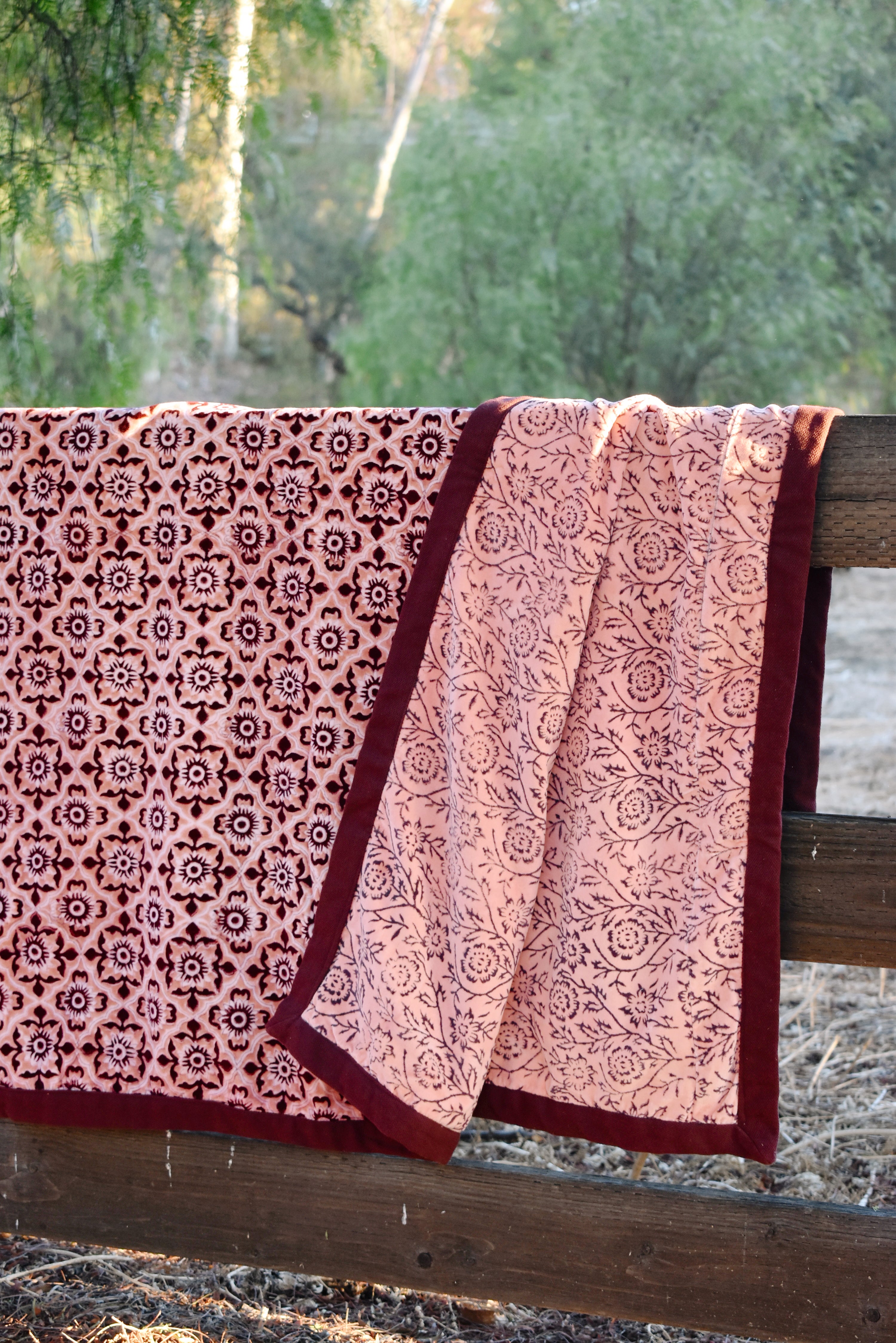 A pink and burgundy floral patterned blanket throw draped over a rustic wooden fence outdoors, with trees in the background. Blanket is open to show reversible print. 