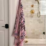 Pink Yarrow block print waffle bath towel hanging from a brass wall hook featuring blue floral vine motifs on a blush pink background