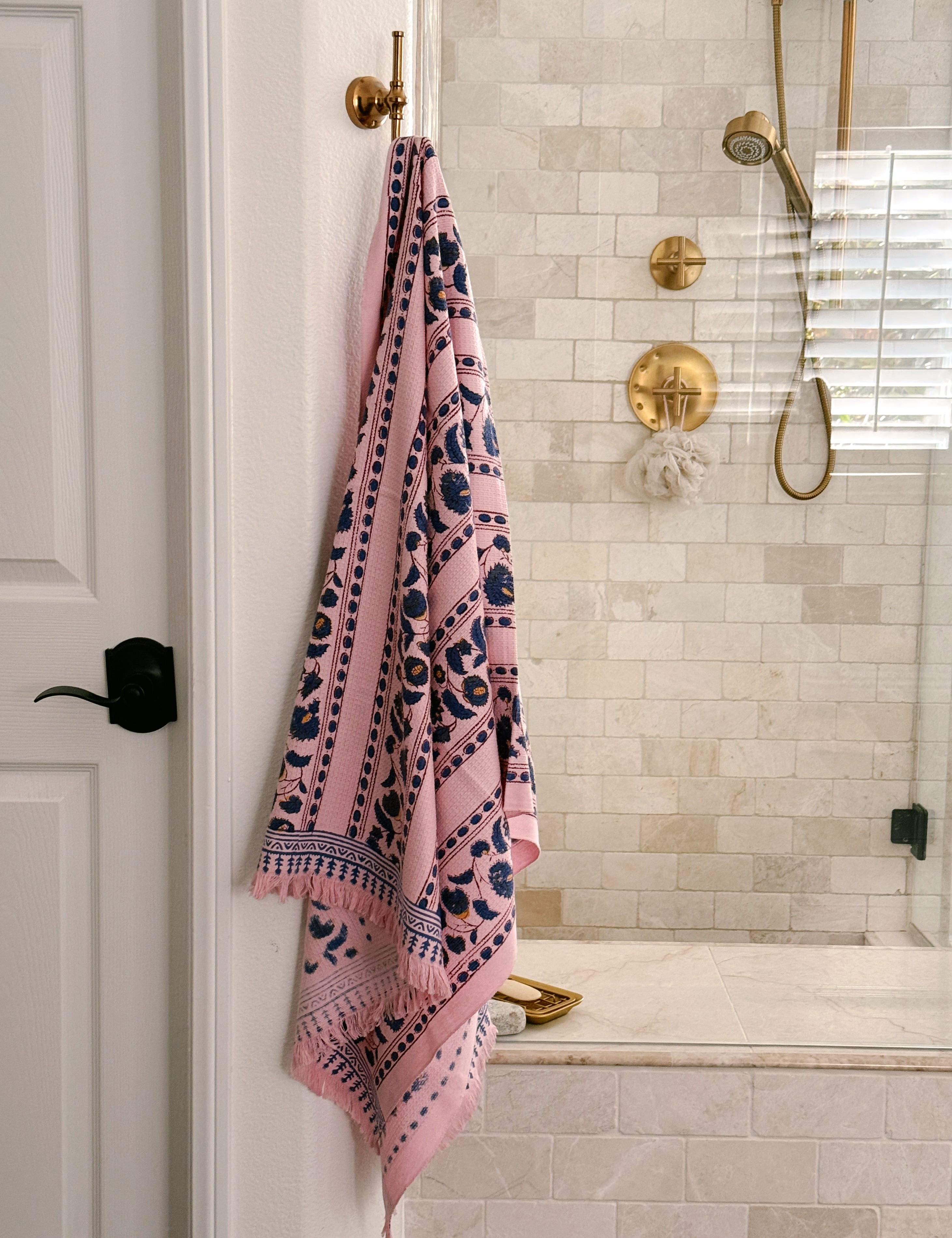 Pink Yarrow block print waffle bath towel hanging from a brass wall hook featuring blue floral vine motifs on a blush pink background