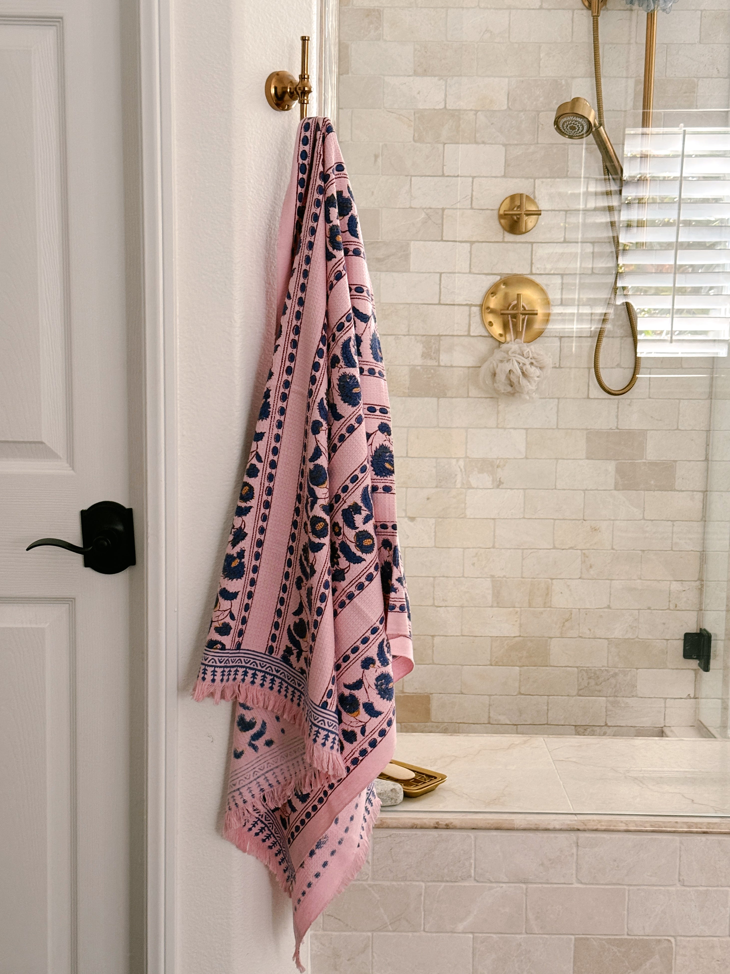 Pink Yarrow block print waffle bath towel hanging from a brass wall hook featuring blue floral vine motifs on a blush pink background