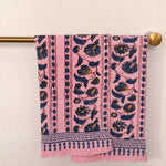 Two pink woven towels with navy blue and mustard yellow floral patterns hangs from a brass rod against a textured off-white wall.