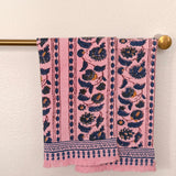 Two pink woven towels with navy blue and mustard yellow floral patterns hangs from a brass rod against a textured off-white wall.