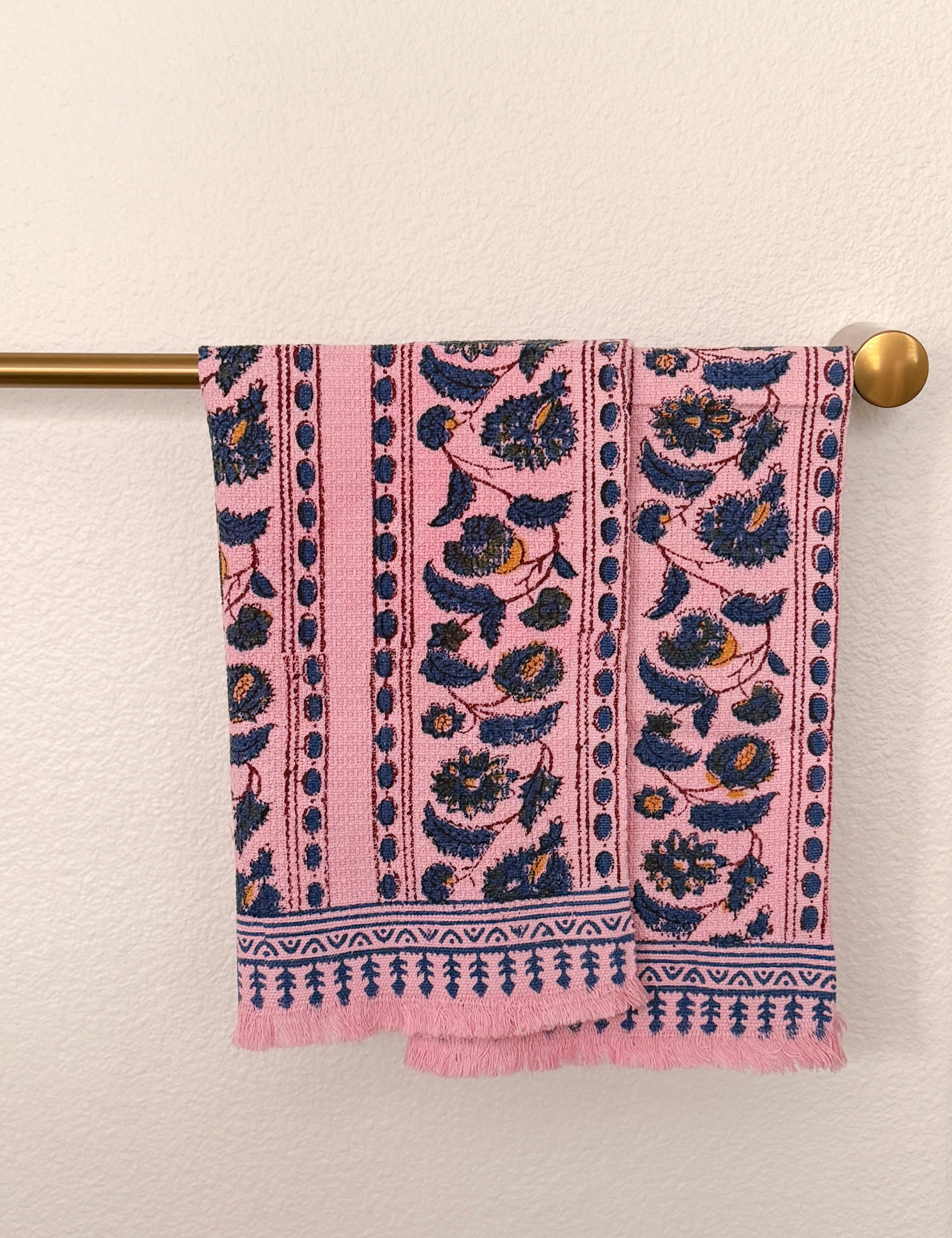 Two pink woven towels with navy blue and mustard yellow floral patterns hangs from a brass rod against a textured off-white wall.
